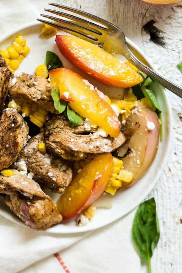 A serving of peach basil balsamic chicken with goat cheese on a small white plate with a fork, on a white linen napkin, on a farmhouse-style surface
