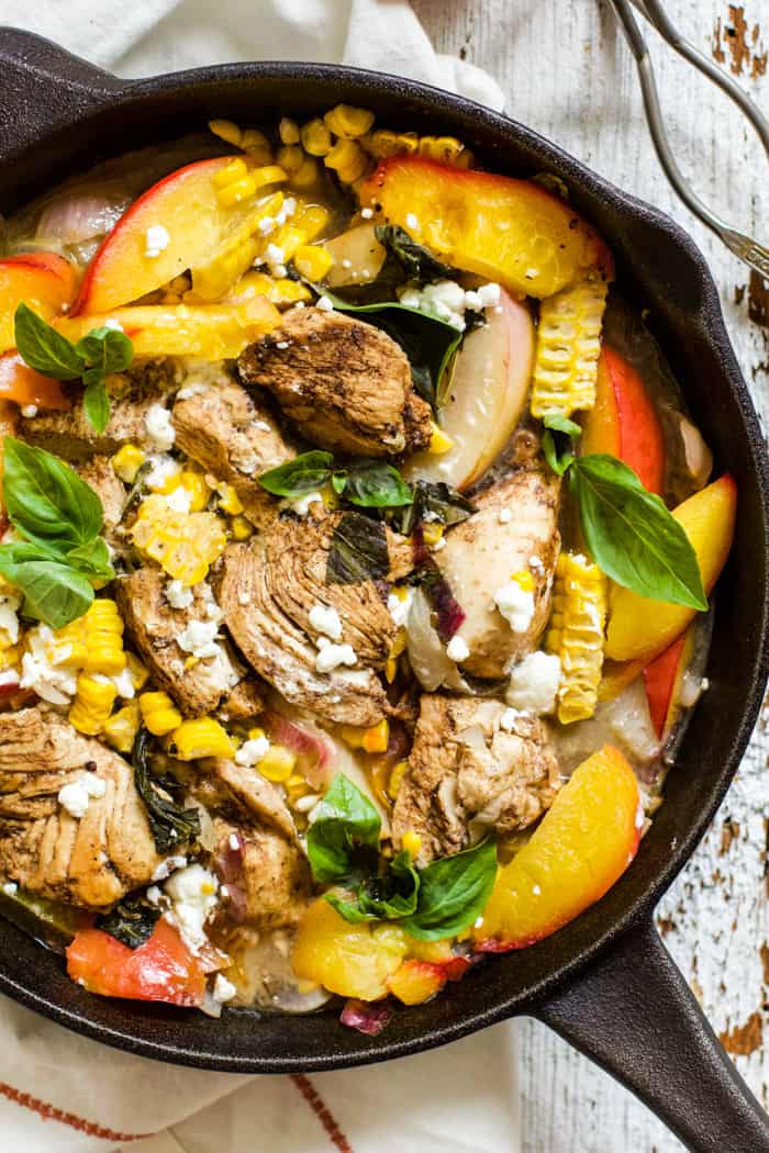 Peach basil balsamic chicken in a cast iron skillet with fresh basil and goat cheese, on top of a white linen napkin on a white farmhouse-style surface