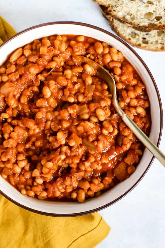 Homemade maple baked beans in beige serving bowl with gold spoon, next to golden-yellow linen, and sourdough slices on the side.