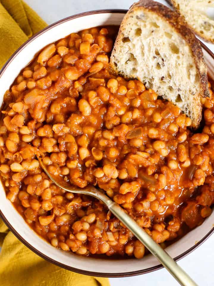 Homemade maple baked beans in beige serving bowl with gold spoon, next to golden-yellow linen, and sourdough dipped in