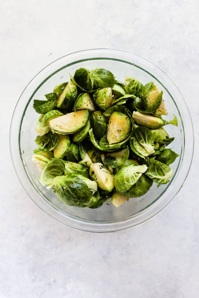Quartered Brussels sprouts tossed in olive oil, maple syrup, salt, and pepper in clear glass bowl