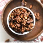 Maple cayenne toasted walnuts in small white bowl on small wooden plate with other walnuts sprinkled around, on white and pink linen