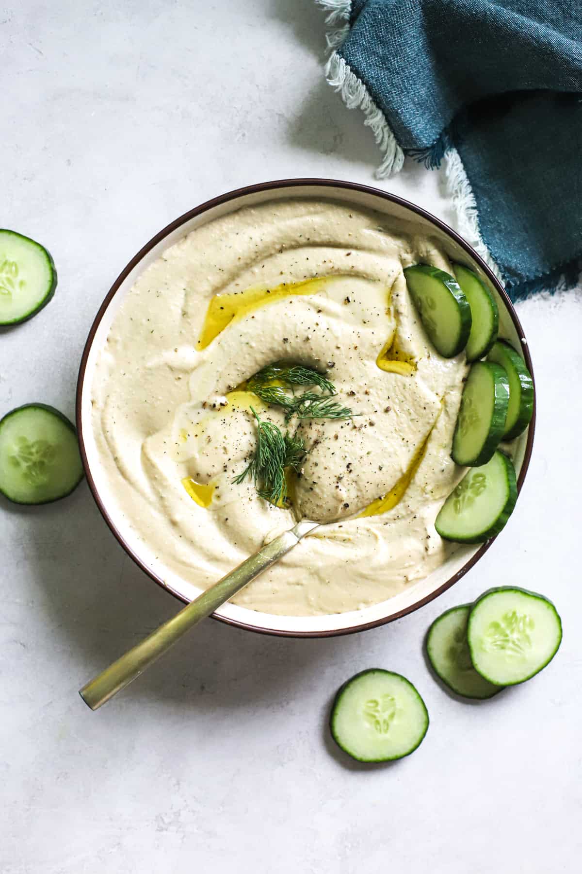 Lemon dill hummus in beige serving dish with olive oil drizzled on top, cucumbers dipped in and on the side, and golden spoon scooped into the hummus.