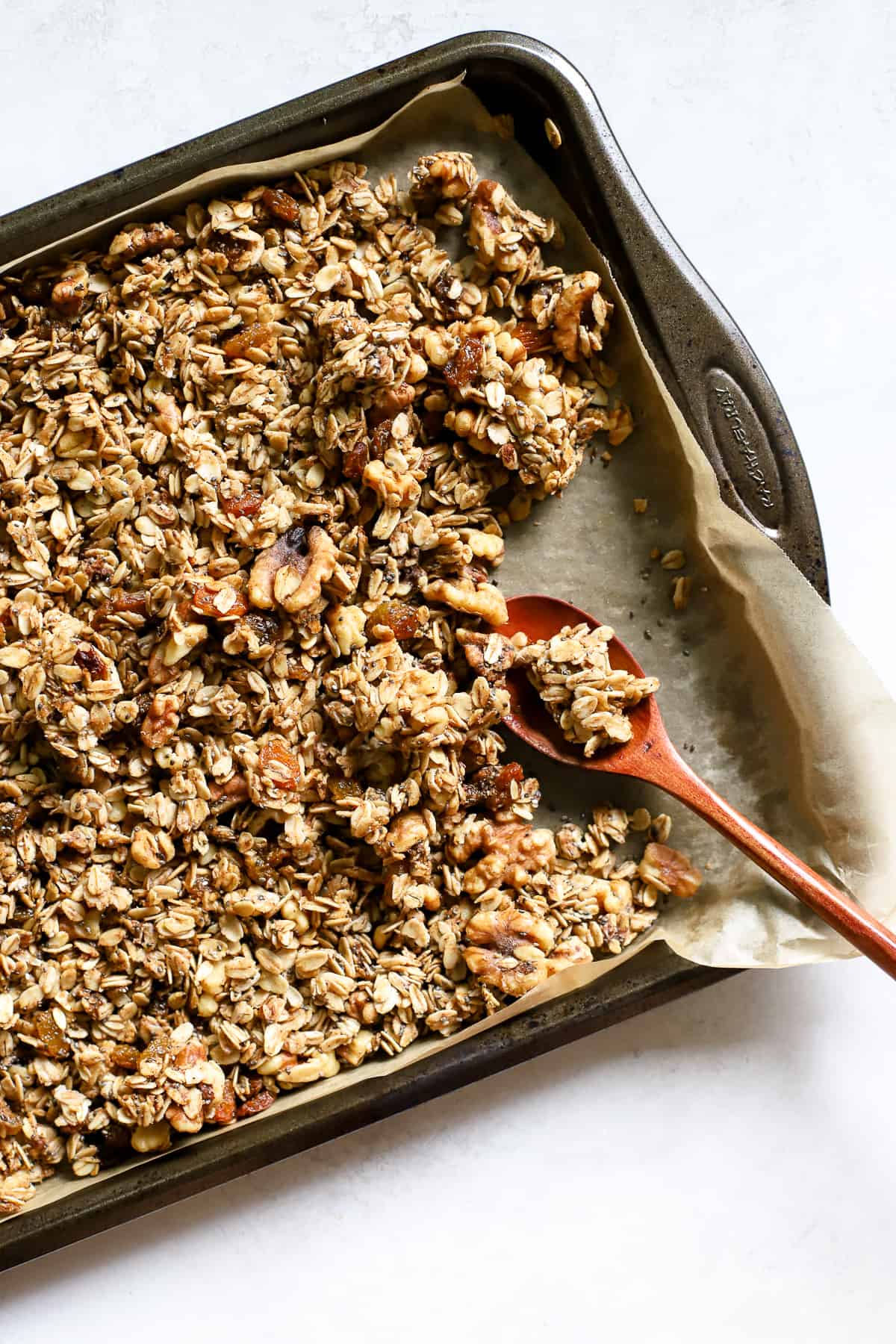 Maple cinnamon walnut granola fully baked, on parchment-lined sheet pan with wooden spoon, and a few clusters broken up