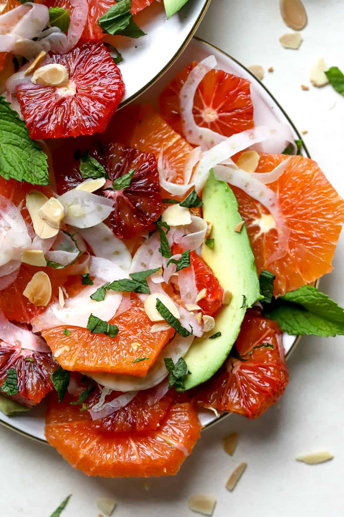 Citrus mint salad with thinly shaved fennel, avocado slices, sliced almonds and chopped mint is served on a cream-colored plate with gold edges. A fork rests on the plate and the plate rests on a beige and cream surface with a few mint leaves and sliced almond crumbs sprinkled around.