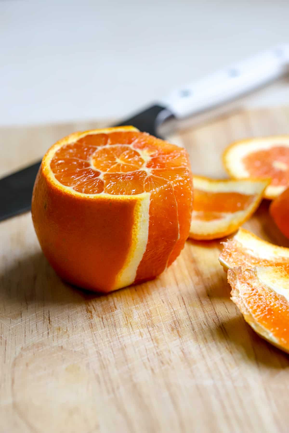 An orange sits on a wooden cutting board with ends sliced off, and some of the peel sliced off to reveal the flesh of the fruit. The orange peels sit on the side and knife sits in the background.