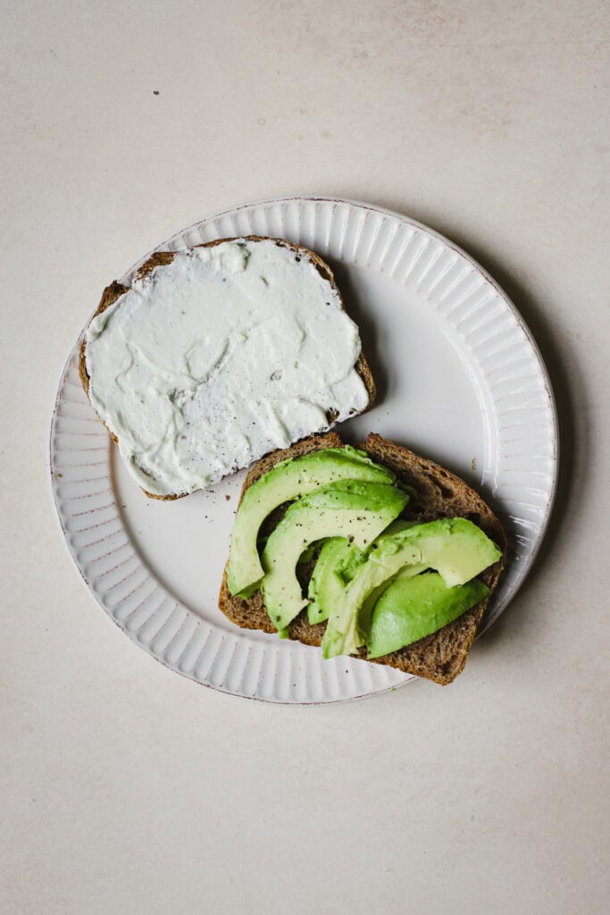 Each sandwich half being assembled with whipped feta and avocado slices.