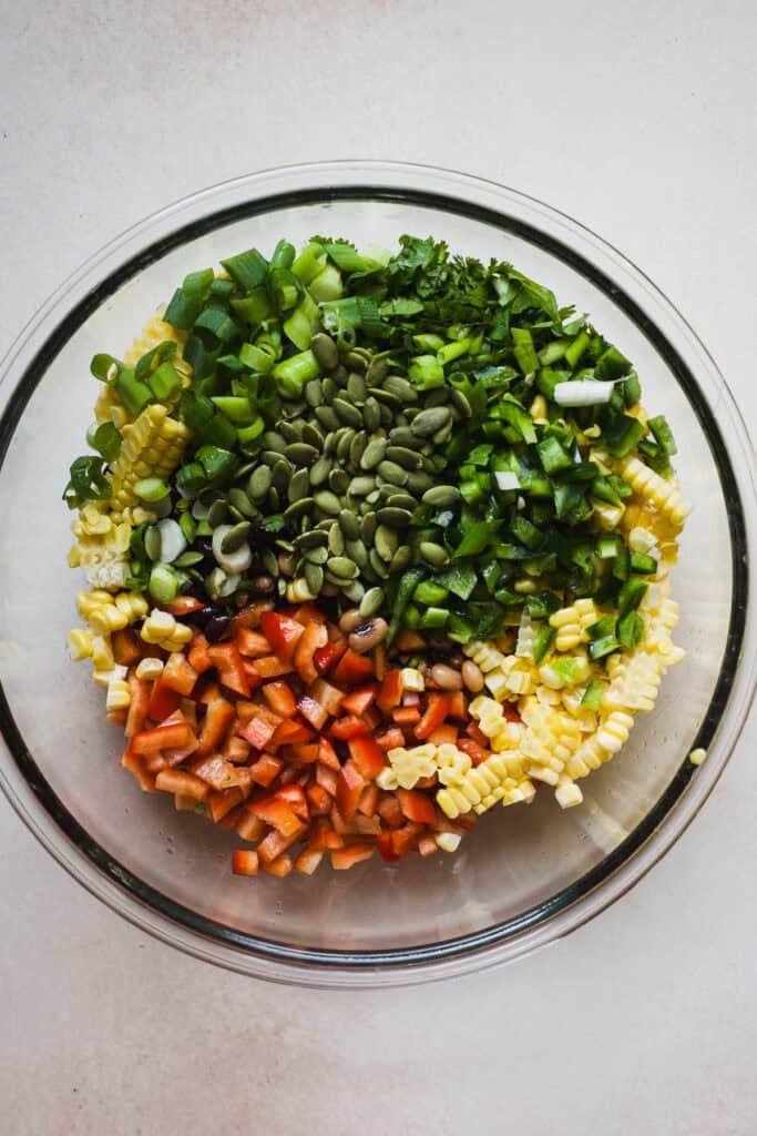 Adding peppers, corn, black beans, black-eyed peas, cilantro, pepitas, green onions, and jalapeños to a bowl.