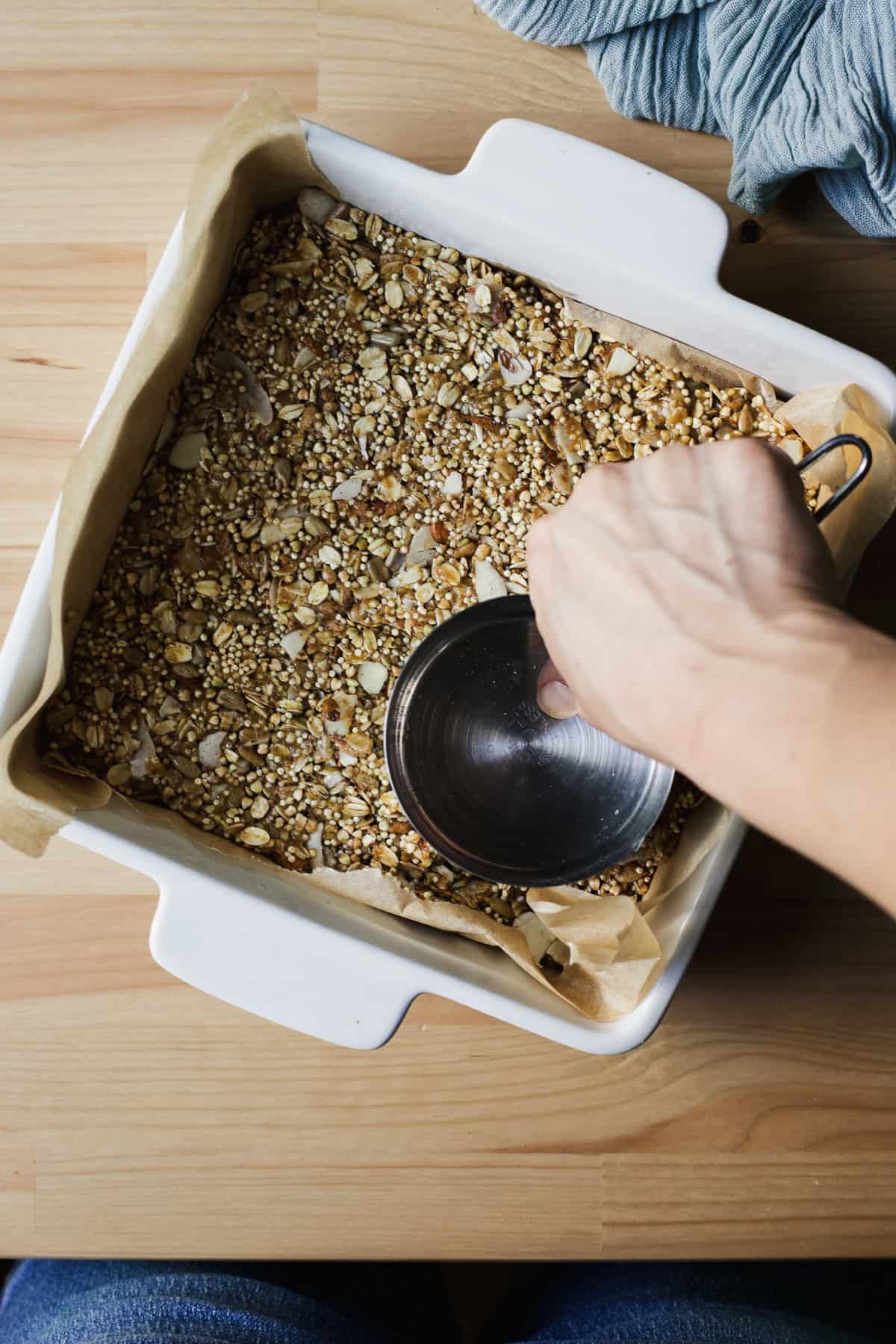 Pressing granola bar mixture into an 8x8-inch ceramic baking dish, using the flat part of a measuring cup.